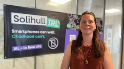 Katie Washbourne stands in front of a banner with the Solihull IRL slogan and the wording underneath says 'Smartphones can wait. Childhood can't'. She has long brown hair and wears a brown sleeveless top with a necklace on top.