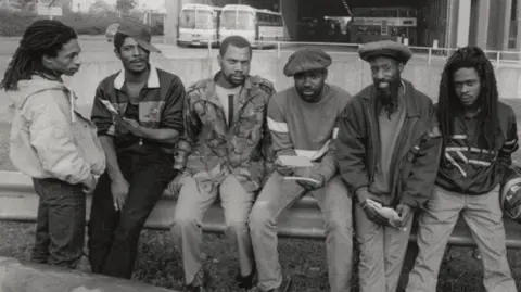 Paul Bingham A group of men in front of a bus station, with of them five sitting down and one standing up. The image is black and white. Three of the men are wearing hats.