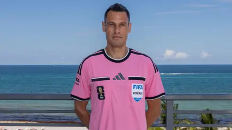 Italian referee Maurizio Mariani standing with hands behind his back wearing a bright pink FIFA Referee shirt stood in front of a white sandy beach with blue sky and sea in the background.