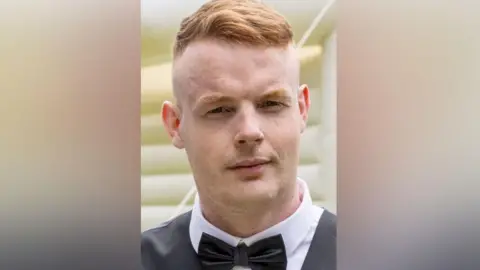 Lewis Bell looks at the camera. He has short, thick ginger hair swept across the top of his head with a very close crop at the sides. He is wearing a white shirt with a black bow tie and grey waistcoat and has a slightly quizzical look.