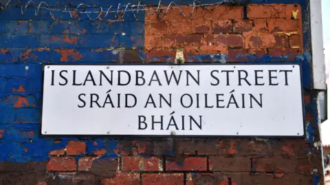 Getty Images A white bilingual street sign in Irish and English. It is on a red brick wall that is partially painted blue. Some barbed wire can be seen on the wall above the sign. 