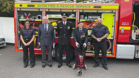 Family Handout Ivor Taylor standing with four members of the fire service in front of a yellow and red fire engine. He is wearing a suit and his war medals, standing up with help from a red walker with wheels. 