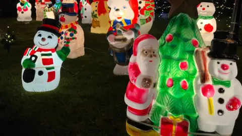 Multiple lit up Christmas figures stand on grass. The display includes various snowmen and a figure of Father Christmas standing next to a Christmas tree.