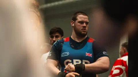 Harry Newby A man with short brown hair in a black top and blue vest with GBR printed on it