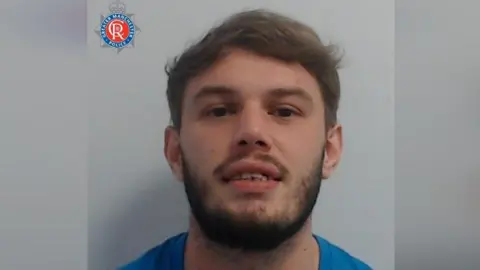 GMP Kyle Camp, with brown hair and a beard and moustache, smirks in a Greater Manchester Police mugshot.