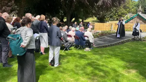 A shot of the backs of a crowd at the service outside under a tree. There are two priests in robes. Some people are sitting and their heads are bowed.