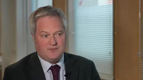 A man in a suit speaks during an interview in an office setting with blinds and wooden doors in the background.