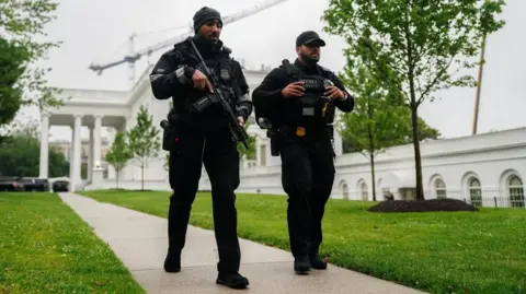 Two Secret Service agents in black uniforms and black hats walk alongside one another on a path outside the White House. One agent is carrying a large gun across his chest. 