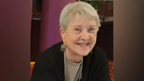 Jenny Collins has short grey hair and is wearing large square earrings, a beige top and a black cardigan. She is smiling at the camera.