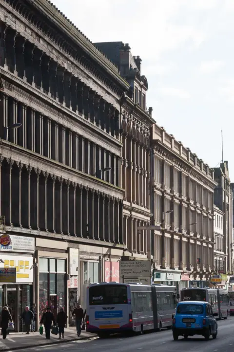 Nick Haynes Side view of the Egyptian Halls, a wide sandstone building with decorative pillars. Busses, a taxi and pedestrians are on the street below.