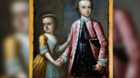 Leeds Museums and Galleries A portrait of a girl and a boy. They are stood together holding hands. The girl is wearing a gold and silver gown while the boy is wearing a pink long jacket with ruffles sleeves and a blue undercoat. He is holding a dress sword.