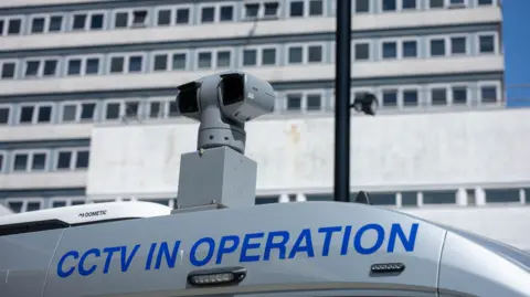 A camera is pictured atop a police van. Only the top part of the van, which is silver in colour, is visible in the image, and the words "CCTV IN OPERATION" are printed in blue text along its side. Beyond the van, there is a large, plain building with rows of windows, like a block of flats.