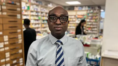 Pharmacist Ade Williams. He is wearing a shirt and a striped blue and white tie. He is looking directly at the camera and behind him are shelves stocked with medicines and pill boxes.