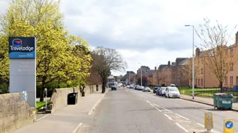 A view of a residential street. A sign for a Travelodge is on one side of the road and the other side has blocks of flats. Cars are parking on the roadside in the distance and streetlamps are on one side of the road.