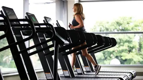 A woman wearing gym gear runs of a treadmill near large glass windows with trees outside. There are several empty treadmills next to her.