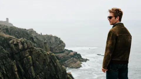 UKTV Damien Molony as Jim Bergerac standing near the edge of a dramatic rocky cliff overlooking the ocean. He's wearing a brown jacket and blue jeans, facing toward the sea. The coastline features rugged rock formations and waves breaking below. 