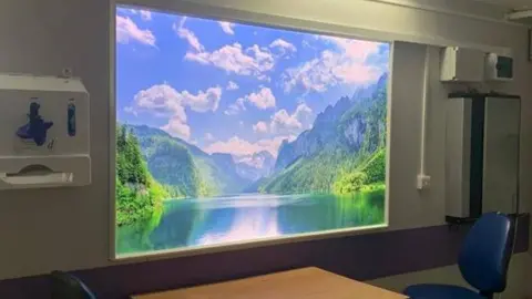 Screen with a shot of a lake which is surrounded by mountains and trees. Light is reflecting on the lake with fluffy clouds visible in the shot.