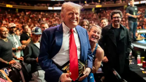 Donald Trump smiles in a vast crowd, holding a UFC belt in front of him