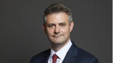 UK PARLIAMENT Man with short grey hair blue blazer white shirt red tie infront of grey background, official portrait 