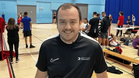 James Robinson from British Dodgeball wearing a black T-shirt. He is standing on a dodgeball court and is smiling into the camera.