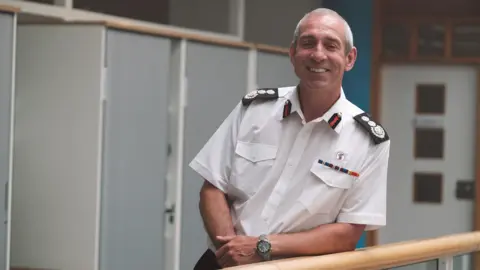 Avon Fire and Rescue Service Simon Shilton. He is pictured leaning his arm on a handrail, looking directly at the camera and smiling. He has short grey hair and is wearing a white shirt. 