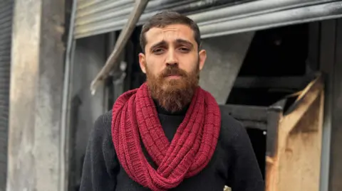 Bashir, a man with dark short hair and a beard looks at the camera wearing a red scarf and a black jumper. He is standing in front of his damaged business in Sandy Row.