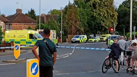 A police cordon of white and blue tape signifying a road closure. There are ambulances and police vehicles beyond it. In the foreground some people have gathered to watch.