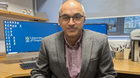 Prof Naveed Sattar sits at his office desk with his computer directly behind him - he looks into the camera and is smiling. He is wearing a textured grey blazer and a purple gingham shirt. He is balding and is wearing thin-rimmed glasses.