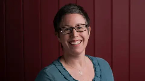 Matt Austin Helen Hartstein, a woman with dark short hair, smiles at the camera. She wears a teal coloured top and glasses and is standing in front of a dark red-coloured wall.