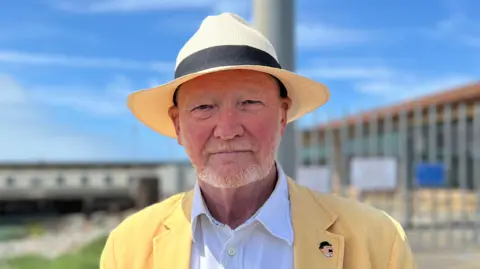 A man with a short grey moustache and beard, wearing a cream hat, looking into the camera. He's wearing a white shirt with the top button undone and a yellow blazer over the top. In the background the sky is blue and his face is shaded by the hat from the sun. There are railings and some buildings behind him.