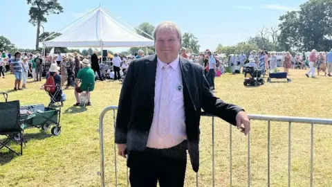 Graham Brown, wearing a black suit with a pale pink shirt and tie, leans on a metal barrier with lots of show-goers behind him. Weather is bright sunshine.