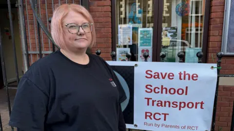 Tina Collins stood next to a sign that says Save the School Transport RCT , run by parents of RCT