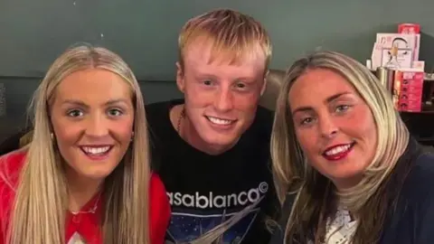 Joshua Perry is sitting in the middle of his mum and sister on a couch. They are all smiling at the camera. 