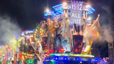 A Ramblers Carnival Club float is being driven down a road in Bridgwater. The float is lit up with blue, yellow and green colours. There are several carnival performers at the front of the float. 