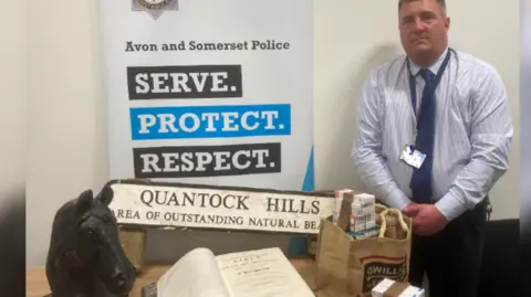 A man in a blue striped shirt, blue tie and dark trousers stands next to a table with various stolen items on it. There is a sculpture of a horse's head, a large hard-backed bible, a sign saying Quantock Hills and a hessian bag full of various packets of medication.