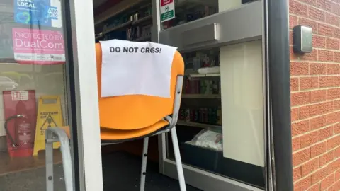 Andrew Turner/BBC An orange chair with a piece of printed paper, stating: "Do not cross!". It is placed in the entrance door of the Wellbeing Pharmacy. Signs can be seen at the window, and the aluminium-framed door is propped open. A red fire extinguisher and a yellow hazard warning sign are visible inside the shop, on the left of the image.