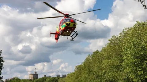 Getty Images A picture of a red helicopter in flight taken from the ground. It is daytime and the sky is blue and full of large white clouds. In the background a castle rises over trees. 