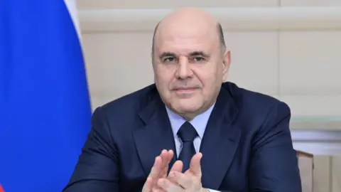 Russian Prime Minister Mikhail Mishustin claps his hands at a ceremony. The blue of a Russian flag falls behind his left shoulder.