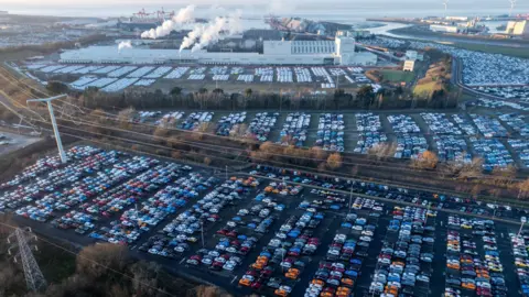 Getty Images Hundreds of new cars are seen parked up in regimented lines at Portbury Dock in Bristol. The picture is taken from the air on a clear day and the Severn Estuary is visible in the background, as are large industrial buildings, some with steam or smoke coming out of them