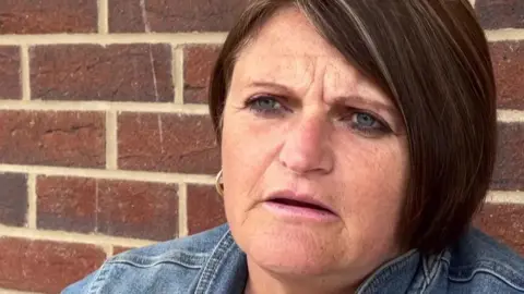 Phil Bodmer/BBC A woman with short brown hair wearing a denim jacket sits against a brick wall.