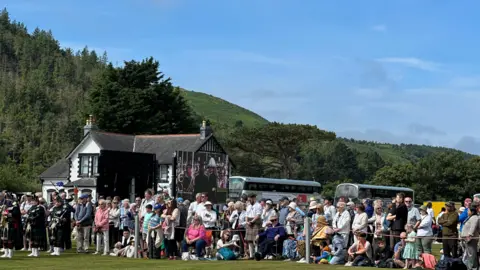 A crowd of people behind a rope barrier watch on with a big screen in the background.