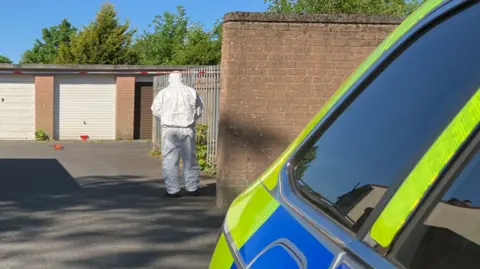 A forensic officer in a white suits walks towards a row of garages with his back to the camera. There is a white and red police tape blocking of entry to the garages which have white and brown roller doors. There is a brown brick wall next to the forensic officer. Beside the camera is a yellow and blue police car.