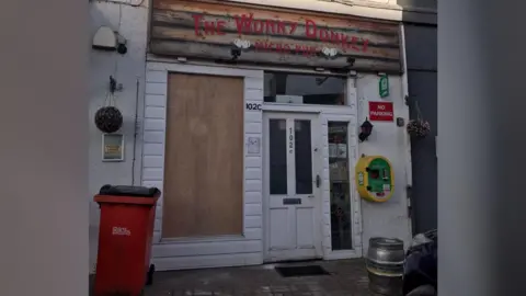 The Wonky Donkey Pub Frontage of the pub building- with a white door and a boarded up window- a red bin in front and a green and yellow defibrillator on the side of the building 