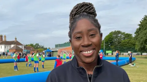 A woman wearing a navy blue coat smiling at the camera. Behind her is a group of children playing football in a field