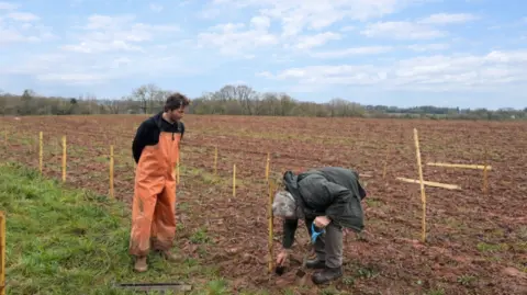A man in orange dungarees and a black top looking at a person in a green coat bending over in a field