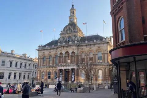 Ipswich Town Hall - the exterior of the ornate sandstone building with two high ceilinged stories and a dome and clock on the top. It overlooks a public square.