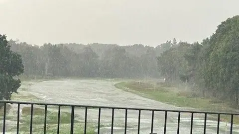 Phil Parker A river of water runs through a fairway on a grassy golf club. There are trees in the background. 