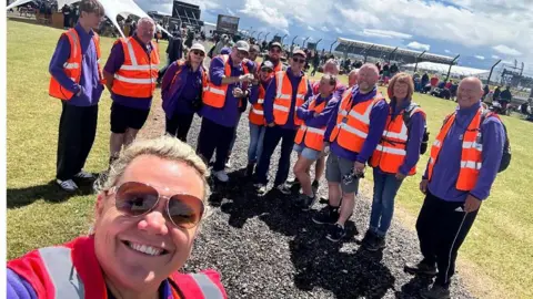 Towcester Community Larder Woman with short blond hair and glasses in a hi-vis stands in front of a row of people in hi-vis at Silverstone