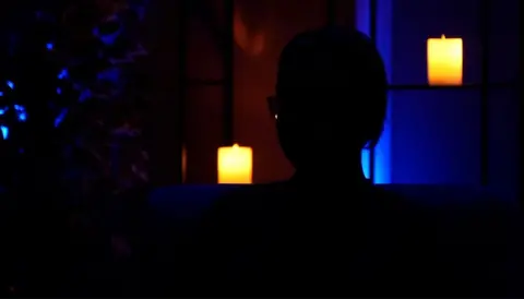 A woman sits in silhouette to protect her anonymity. She is sitting in front of a blue backdrop with a shelf with electric powered candles. 
