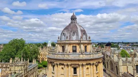 Angela SATURDAY - The Radcliffe Camera is a round yellow stone building with a grey roof in the centre of Oxford, behind you can Oxford city centre and a green tree. In the distance you can see trees on the horizon. The sky is blue with white and grey clouds.